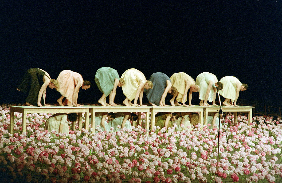 Scena ze spektaklu Nelken ukazująca osiem tancerek w pełnym ukłonie, w widoku z profilu, stojących na długim stole. Na pierwszym planie setki biało-różowych goździków.