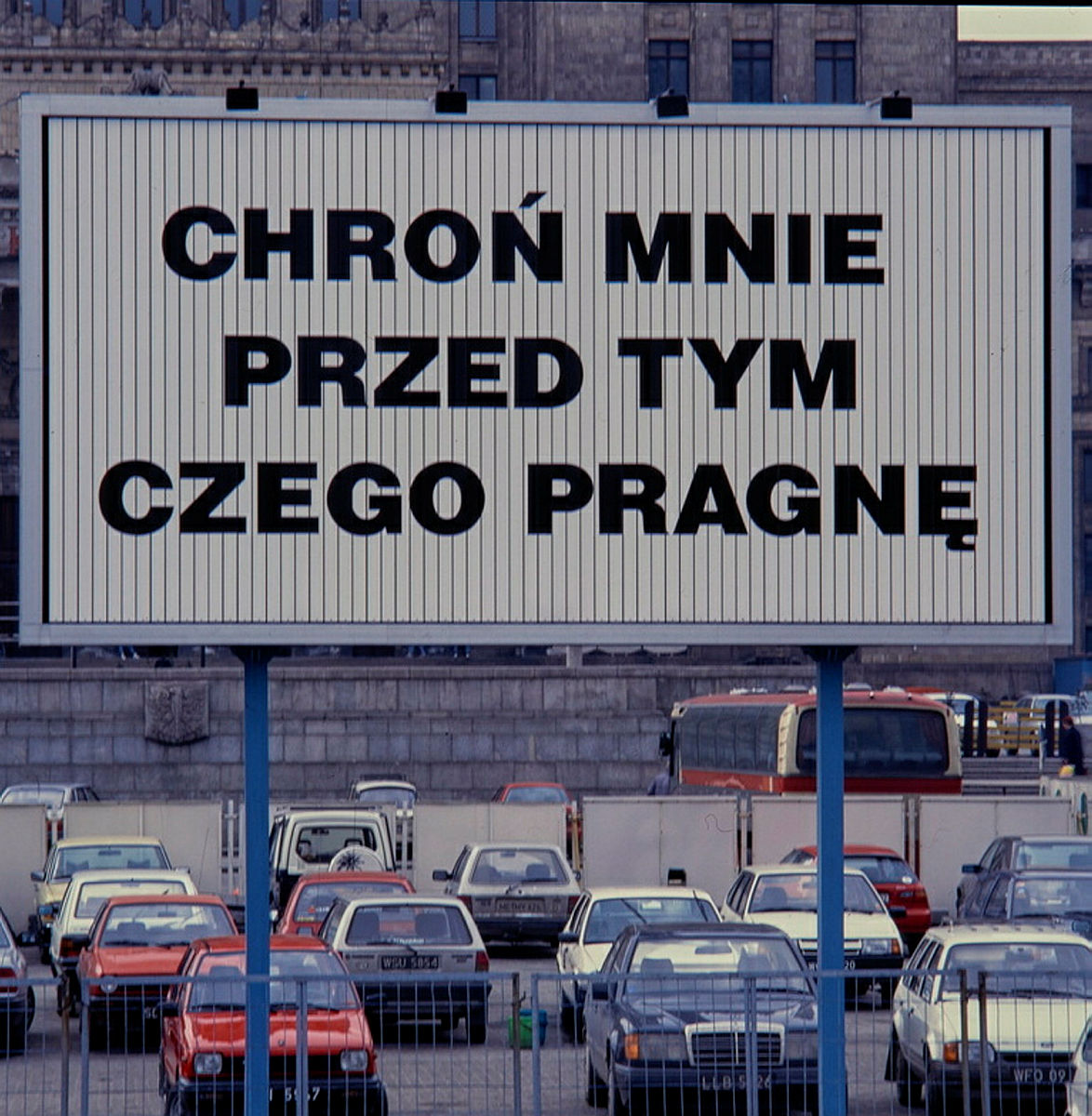 W centralnym polu fotografii znajduje się praca artystki Jenny Holzer- wielkoformatowy billboard z hasłem: CHROŃ MNIE PRZED TYM CZEGO PRAGNĘ. W otoczeniu billboardu znajdują się samochody. 