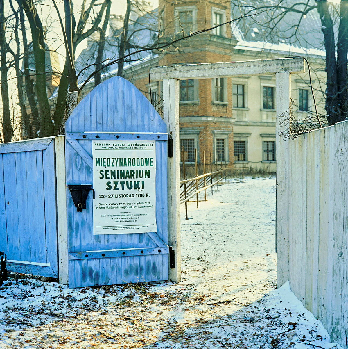 Drewniany płot okalający remontowany zamek ujazdowski w pejzażu zimowym. Na płocie czarno biały plakat informujący o Międzynarodowym Seminarium Sztuki.